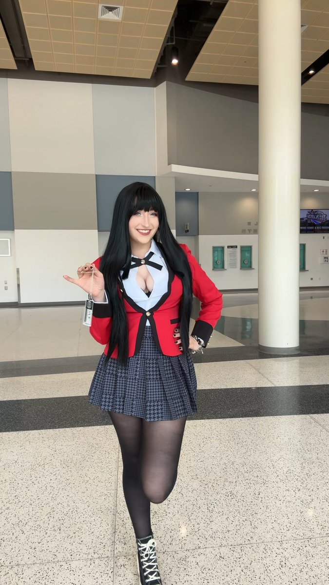 A woman with long black hair wearing a red blazer, plaid skirt, and black tights poses in what appears to be a convention center hallway.