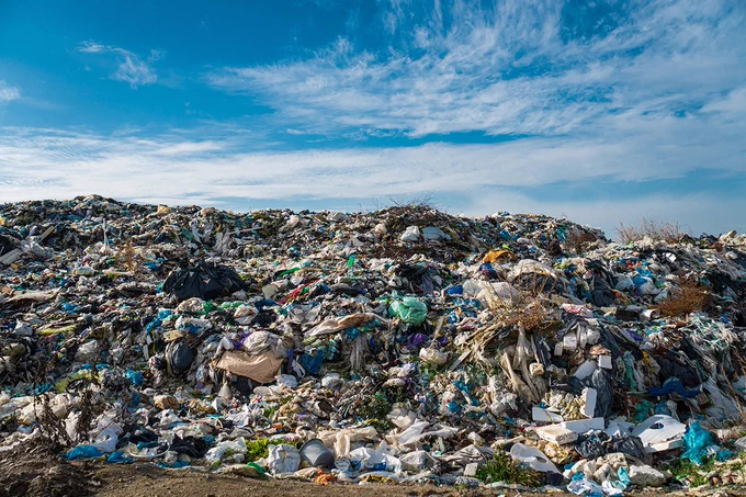 A vast and towering landscape-sized mound of mixed garbage and debris, predominantly plastics, fills the lower two-thirds of the image. The enormous pile of refuse sits under a bright blue sky with scattered white clouds, illustrating significant waste accumulation.