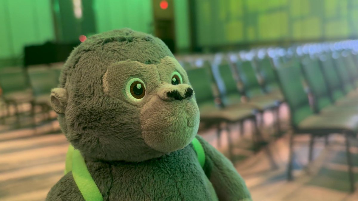 A close-up of a stuffed monkey toy with a green strap, photographed in what appears to be an airport waiting area with rows of empty seats in the background.
