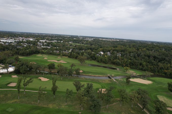 An aerial view of a lush green golf course featuring sand traps, water features, and cart paths, surrounded by trees and residential areas under a cloudy sky.
