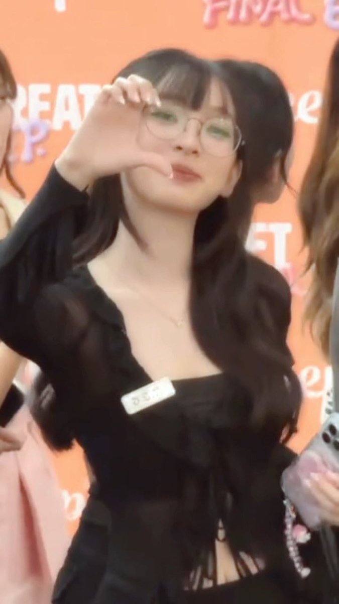 A young woman with long dark hair, wearing glasses and a black outfit, makes a hand gesture in front of an orange backdrop with partial 'FINAL' and 'CREAT' text visible.