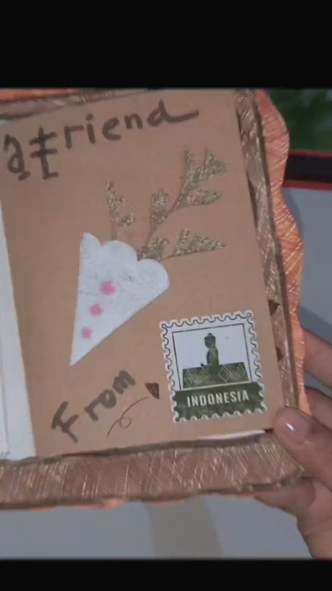 A handmade greeting card held in someone's hand, featuring a brown paper cover with the text "atriena", "from", a white heart with pink dots, dried flowers, and an "INDONESIA" postage stamp.