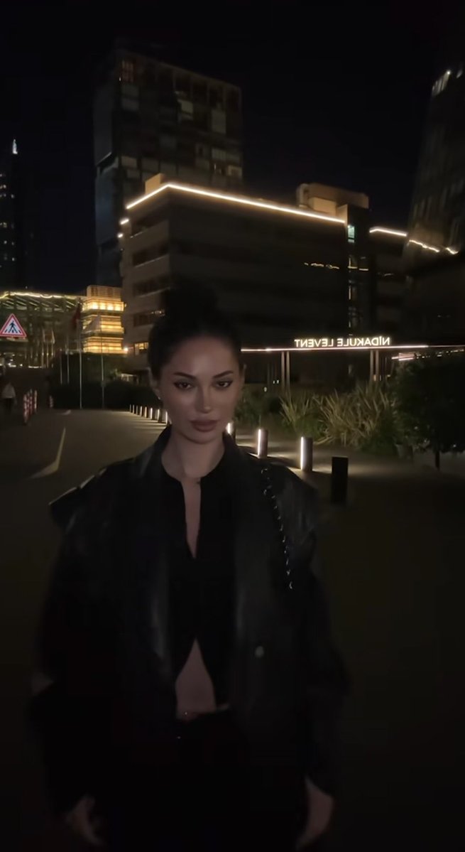 A woman with dark hair in a bun wearing a black leather jacket stands outside at night, with the illuminated "HIDEAUX LE 3 F EVENT" sign on a building behind her.