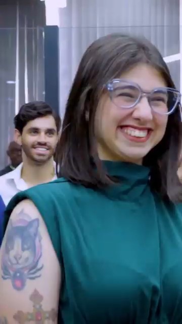 A smiling young woman wearing glasses and a dark green sleeveless top stands in an elevator, with a man visible behind her; she has a colorful cat and cross tattoo on her upper arm.
