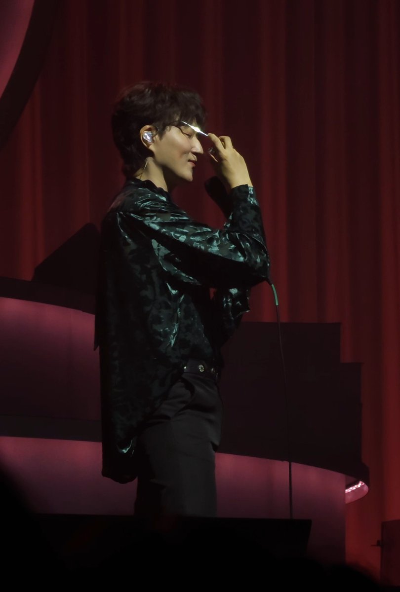 A male performer in a dark patterned shirt and glasses smiles while holding a microphone on stage in front of dark red curtains.
