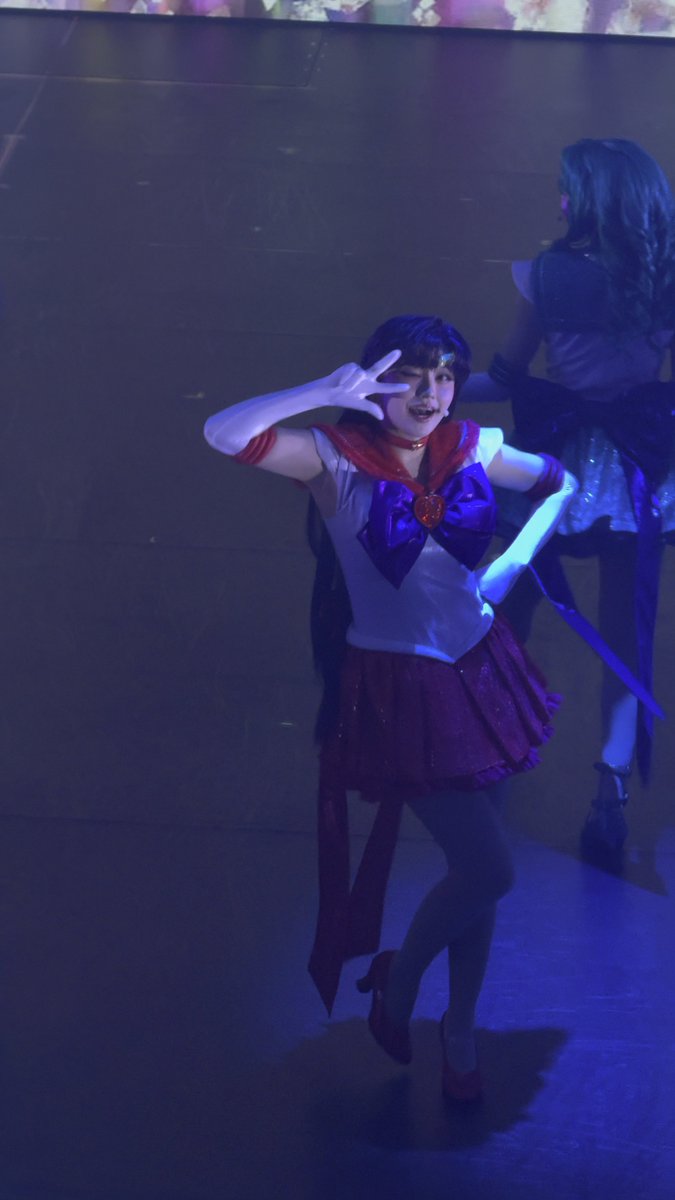 A performer in a Sailor Moon-inspired costume poses on a stage bathed in blue lighting, making a peace sign with her hand while another performer stands in the background.