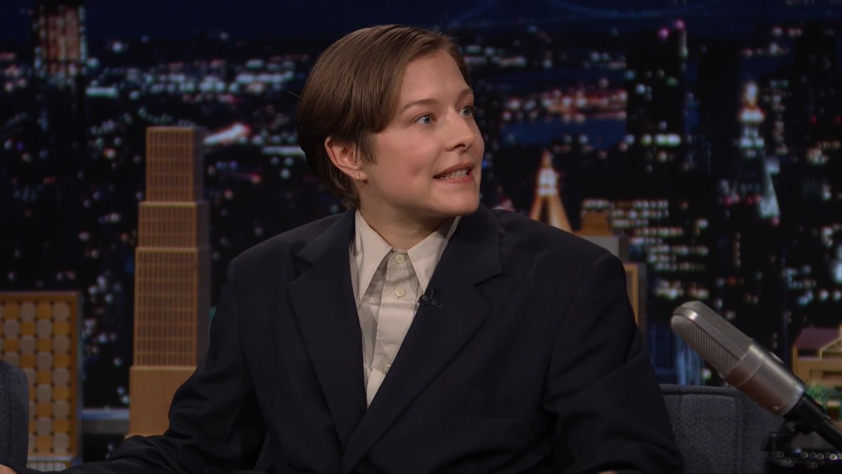 Emma D'Arcy sits on a talk show set wearing a dark blazer over a cream button-up shirt, with short brown hair and a lavalier microphone, appearing engaged in conversation with a silver microphone positioned in the foreground and a city skyline backdrop behind them.