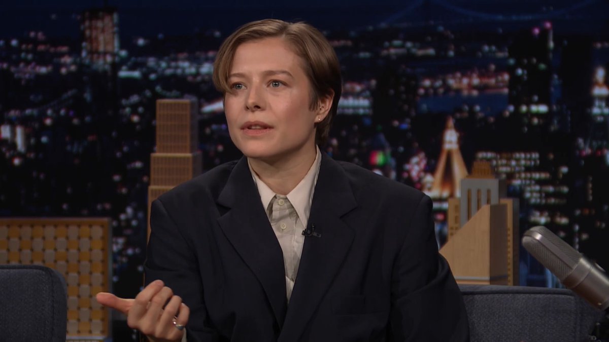 Emma D'Arcy appears on The Tonight Show with Jimmy Fallon, wearing a dark blazer over a white shirt and gesturing while discussing their new film "Digger," with a nighttime cityscape backdrop behind them.