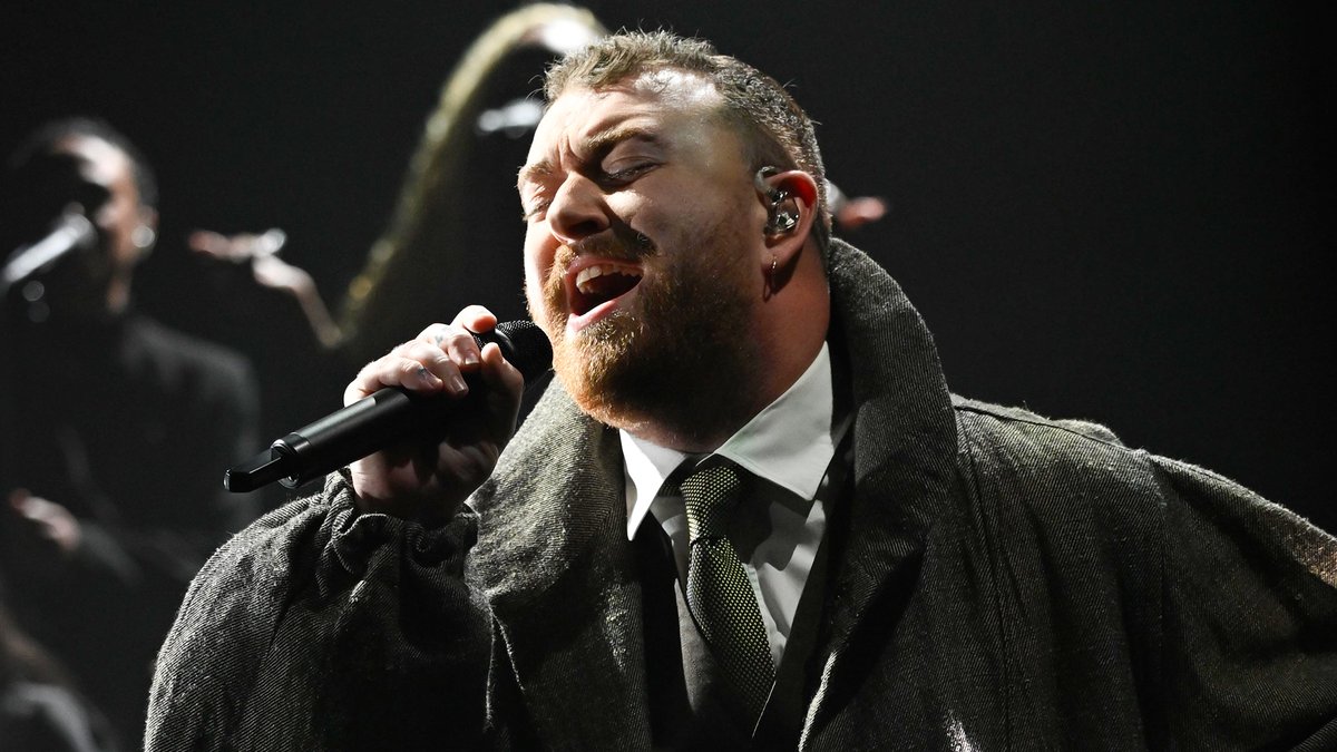 Sam Smith passionately sings into a microphone during a live performance of "Oh Mother" on The Tonight Show, dressed in a textured gray coat with a white shirt and dark tie, while a saxophonist performs in the dimly lit background.