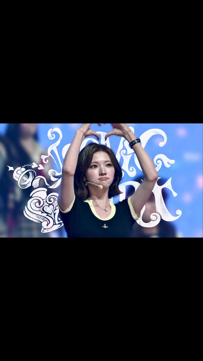 A young woman performing on stage with her hands raised above her head forming a shape, wearing a black and yellow shirt with a headset, against a blue background with decorative graphics.