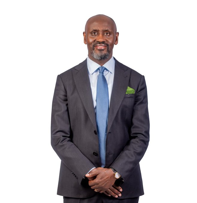 A professional portrait of a smiling bald man with a gray beard wearing a dark suit, light blue shirt, blue tie, and green pocket square.