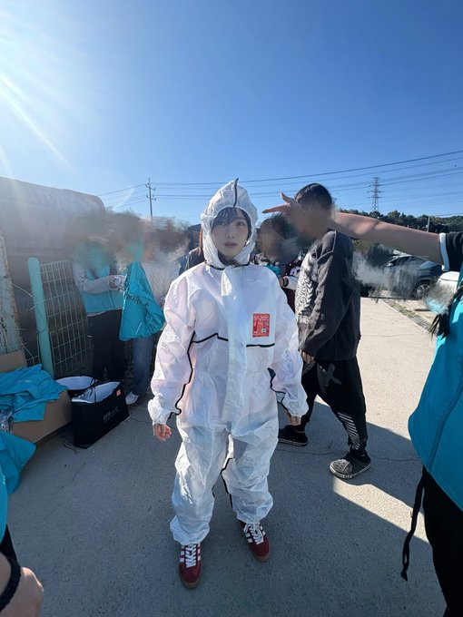A person in a white protective suit stands in an outdoor area, surrounded by others, with smoke visible in the background under a clear blue sky.