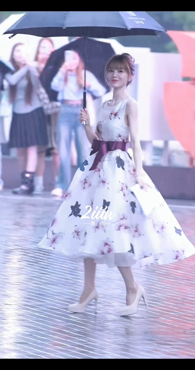 A woman in a white floral dress with a purple bow waist stands under a black umbrella on a wet stage, with blurred figures in the background and a watermark reading '紫之靛'.