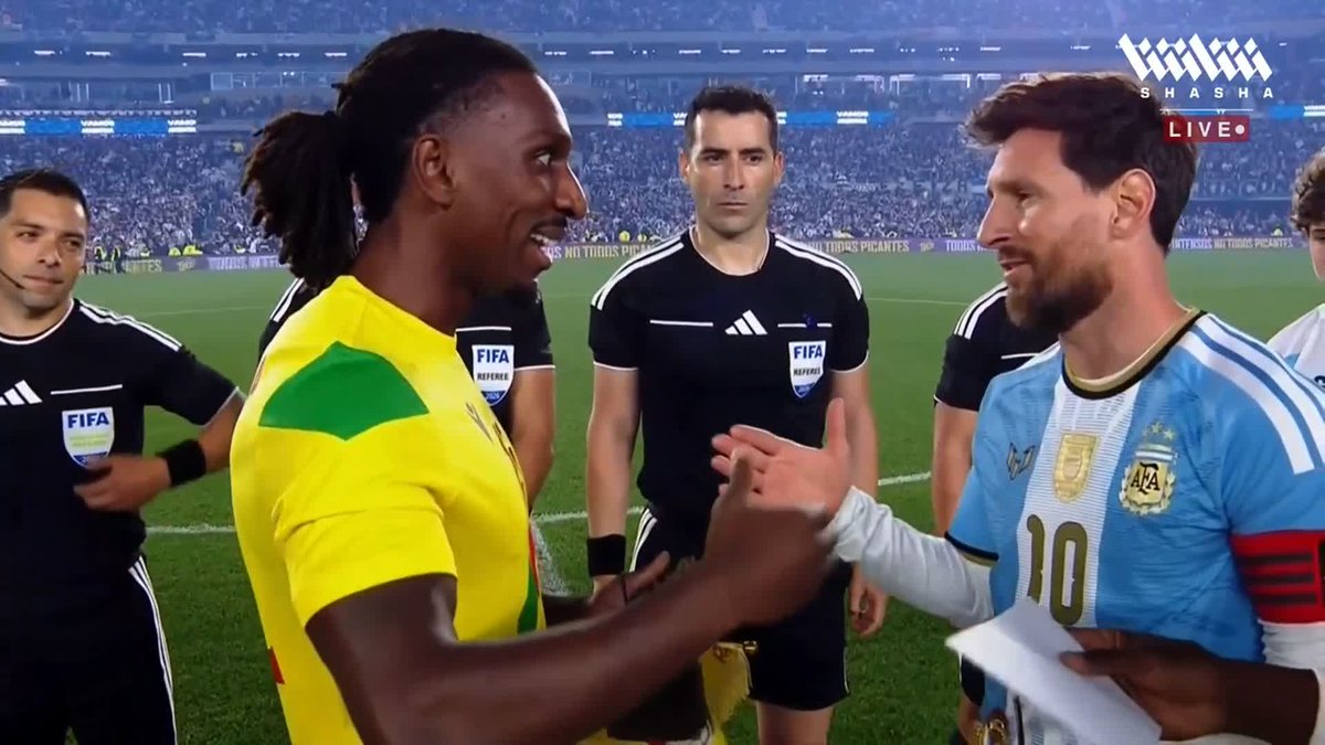 Lionel Messi, wearing the Argentina #10 jersey, shakes hands with a Benin player in a yellow and green kit on the pitch, with FIFA referees standing nearby. The "Shasha Live" watermark is visible in the corner, indicating this moment from Messi's retirement match is being broadcast on the Shasha app, with the Benin player appearing to present a commemorative gift.