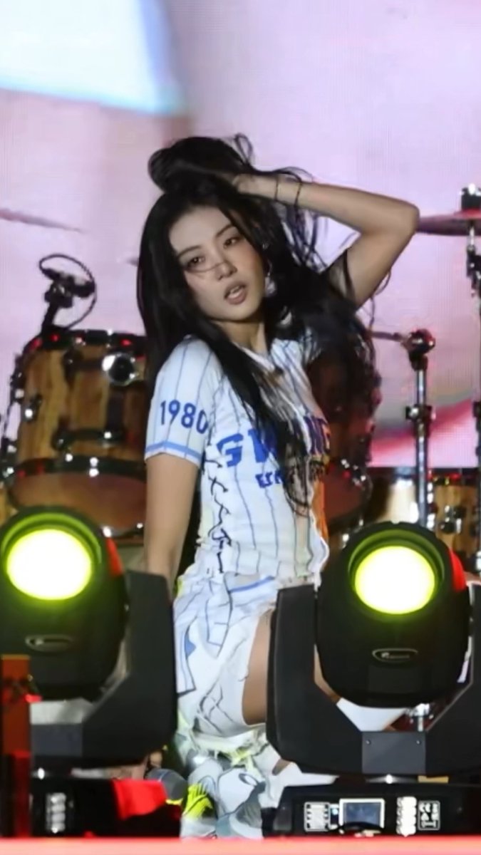 A female performer with long dark hair performs on stage in a white jersey marked '1980' and '53', with green-lit stage lights in the foreground and drums behind her.