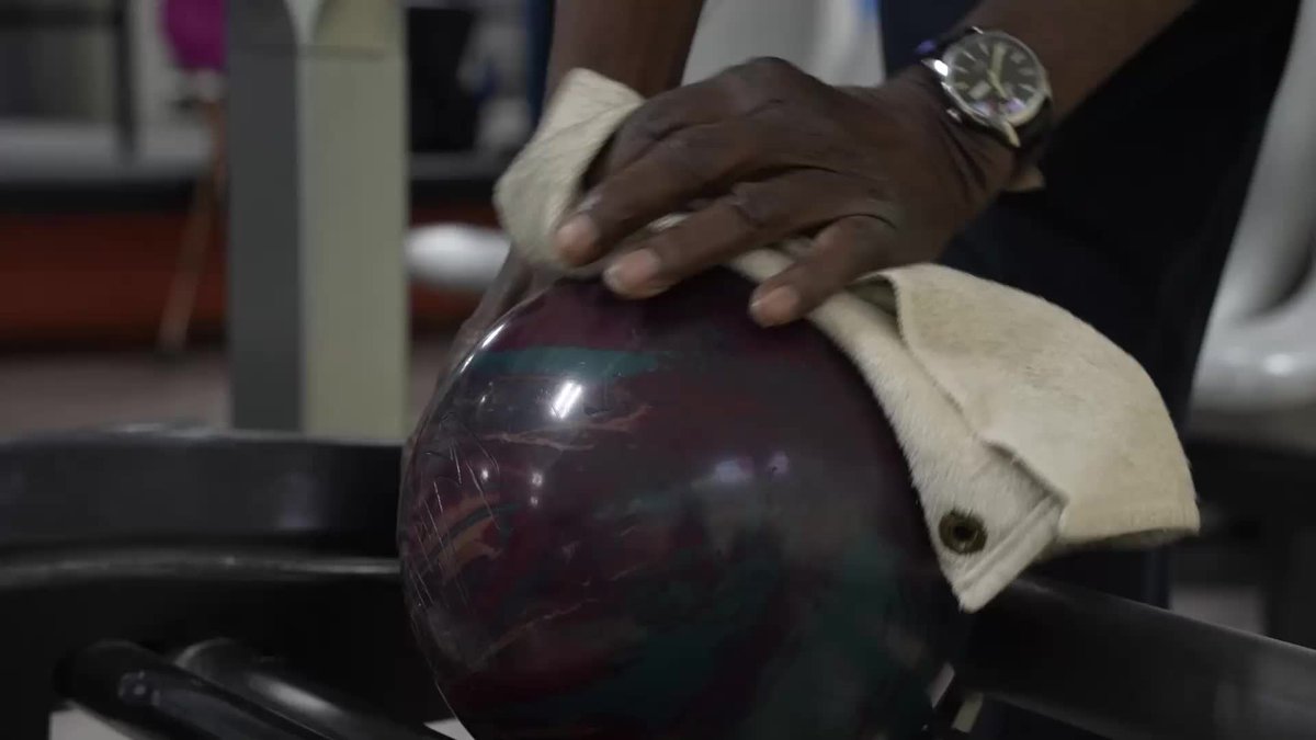 A pair of hands, one wearing a watch and cradled in a white towel, grips a marbled purple and green bowling ball as a 100-year-old bowler prepares for his next frame, defying his age with every throw.