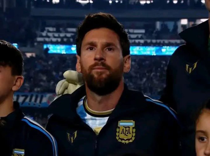 Lionel Messi wearing an AFA (Argentine Football Association) tracksuit jacket while standing on a stadium field, appearing somber with children around him.
