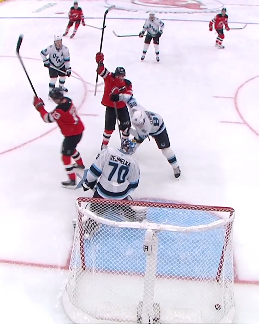 A New Jersey Devils player in a red jersey celebrates with his stick raised after scoring a goal against the Seattle Kraken, with goaltender Vejmelka (#70) looking dejected in the net. The name "VEJMELKA" and number "70" are visible on the goalie's jersey, capturing the Devils' triumphant moment.