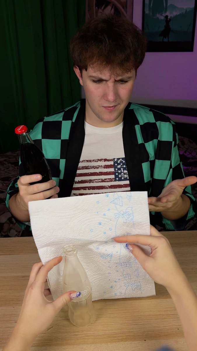 A young man in a checkered jacket and American flag t-shirt sits at a table holding a bottle of dark soda, looking confused as hands in the foreground hold a napkin and an empty bottle toward him.