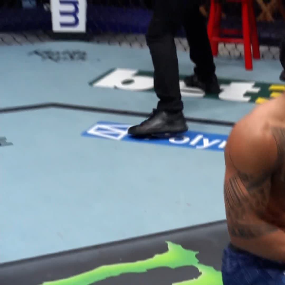 A person's black shoes and legs are visible on the mat of an MMA octagon, with "OLYM" sponsor text on the floor and a green Monster Energy logo in the foreground, while a victorious Brazilian fighter stands by following a first-round knockout victory on Dana White's Contender Series.