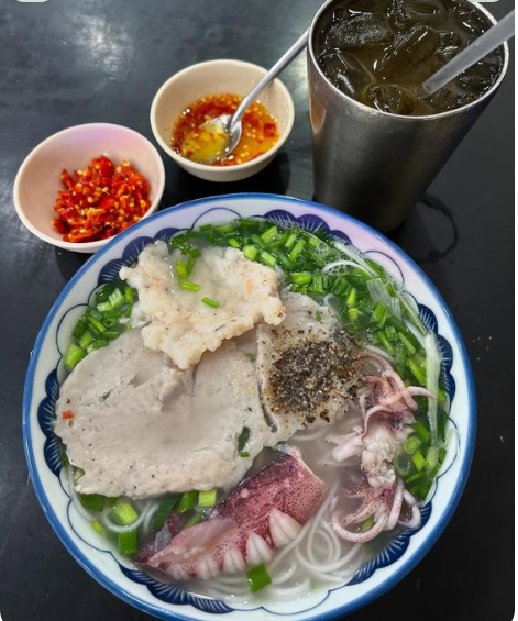 A bowl of Vietnamese noodle soup features pork patties, squid, shrimp, chives, and noodles in a clear broth, accompanied by a side of chopped red chilies, chili sauce, and a metal cup of iced tea.