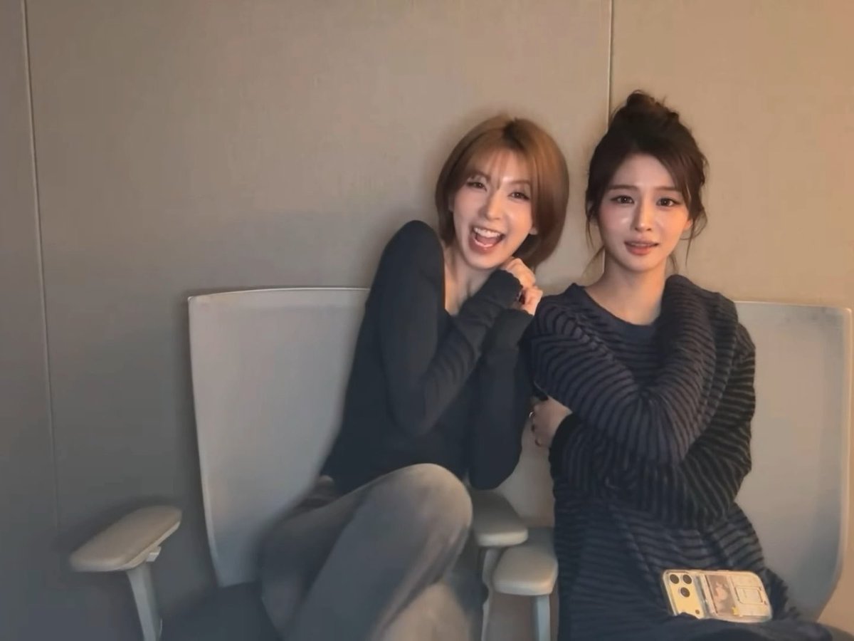 Two young women sitting on a light-colored bench; one has short brown hair and is smiling broadly, while the other has dark hair in a bun and wears a dark striped sweater, holding a phone with a decorative case.