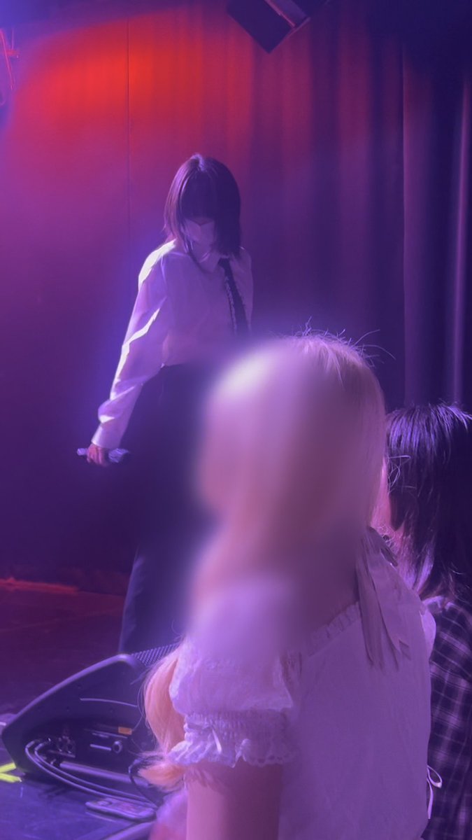A performer with short dark hair, wearing a white shirt and dark tie, stands on stage under purple and red lighting, with blurred figures in the foreground.