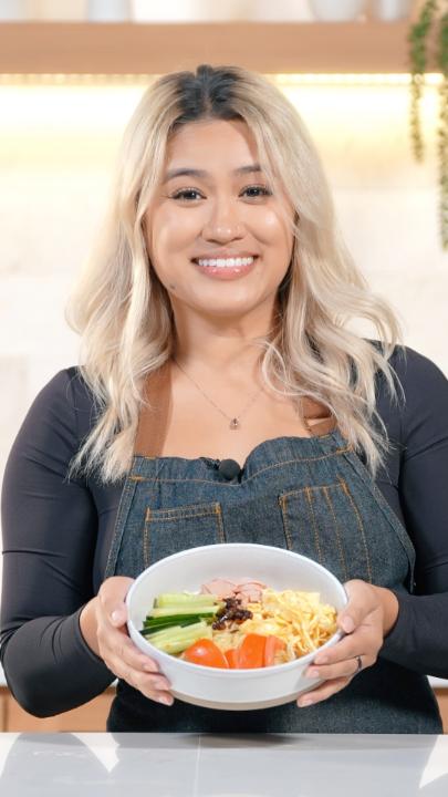 A smiling woman with long blonde hair, wearing a denim apron over a black top, holds a white bowl filled with colorful noodle ingredients including cucumber, tomato, egg, and vegetables in front of a marble kitchen backsplash.

The image relates to the post by presenting Keele, the cheerful host showcasing a prepared noodle dish to promote the Tales of Eternia Remastered launch on October 16th.