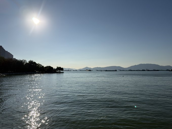 A serene photograph of a wide expanse of water reflecting bright sunlight, with silhouettes of distant mountains, cargo ships, and a city skyline along the horizon.