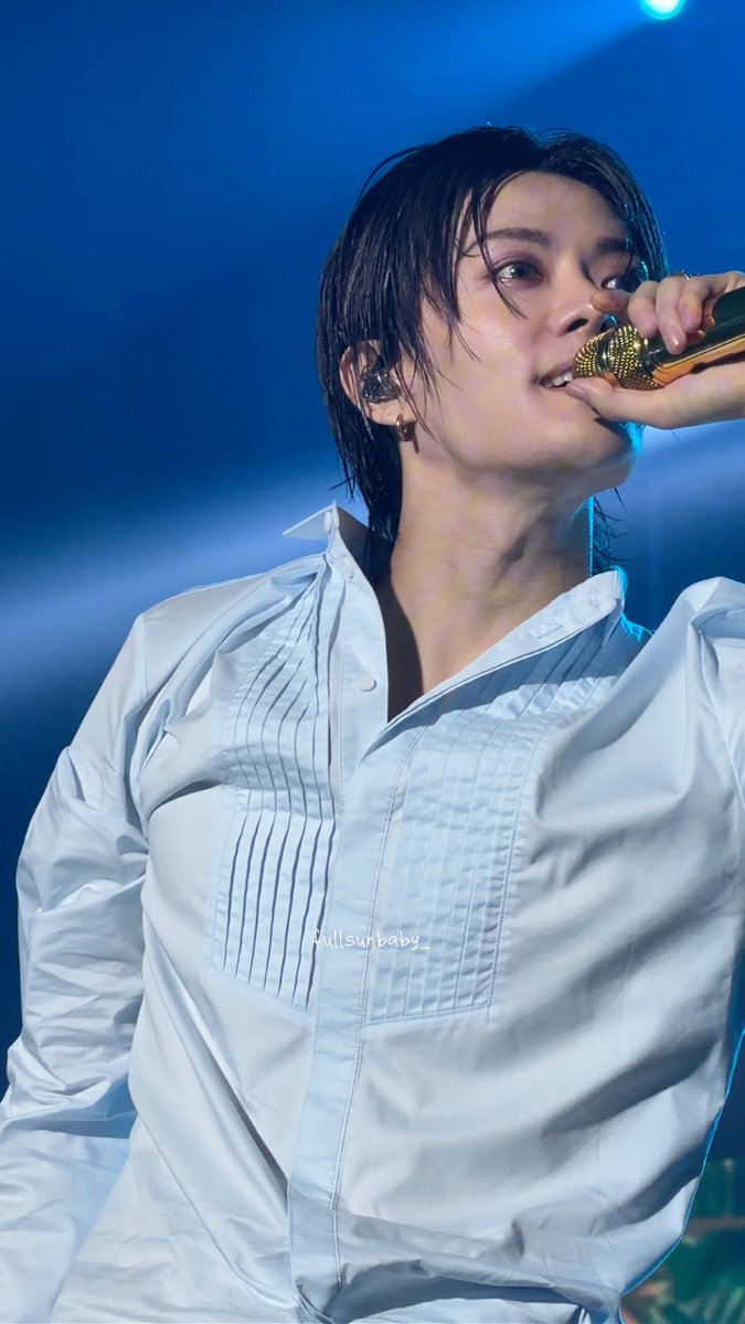 A person performs passionately into a gold microphone under blue stage lighting, wearing a crisp white pleated shirt with damp hair framing their face, alongside the hashtag "NCT127_5TH_NEOCITY_THE_REDLINE#THE_REDLINEinJakarta" indicating this is NCT 127 member Yuta during the Jakarta leg of their tour.