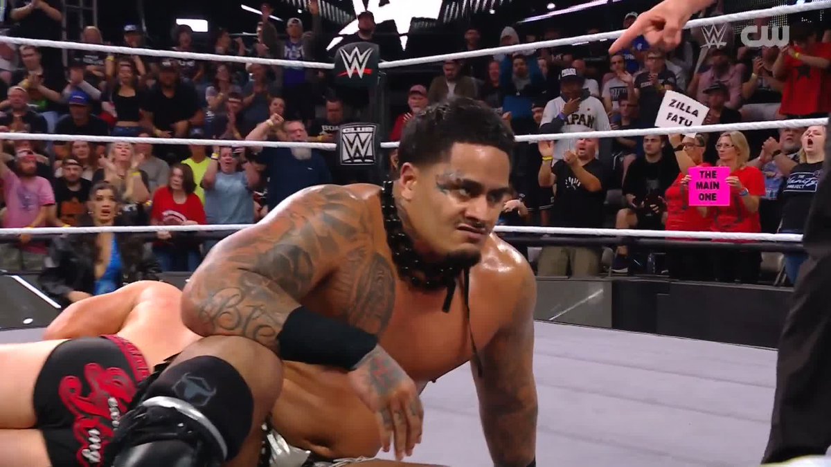 A tattooed wrestler kneels inside the WWE ring on The CW, with fans holding signs reading "THE MAIN ONE" and "ZILLA FATU," visually confirming the post celebrating Zilla Fatu's victory.