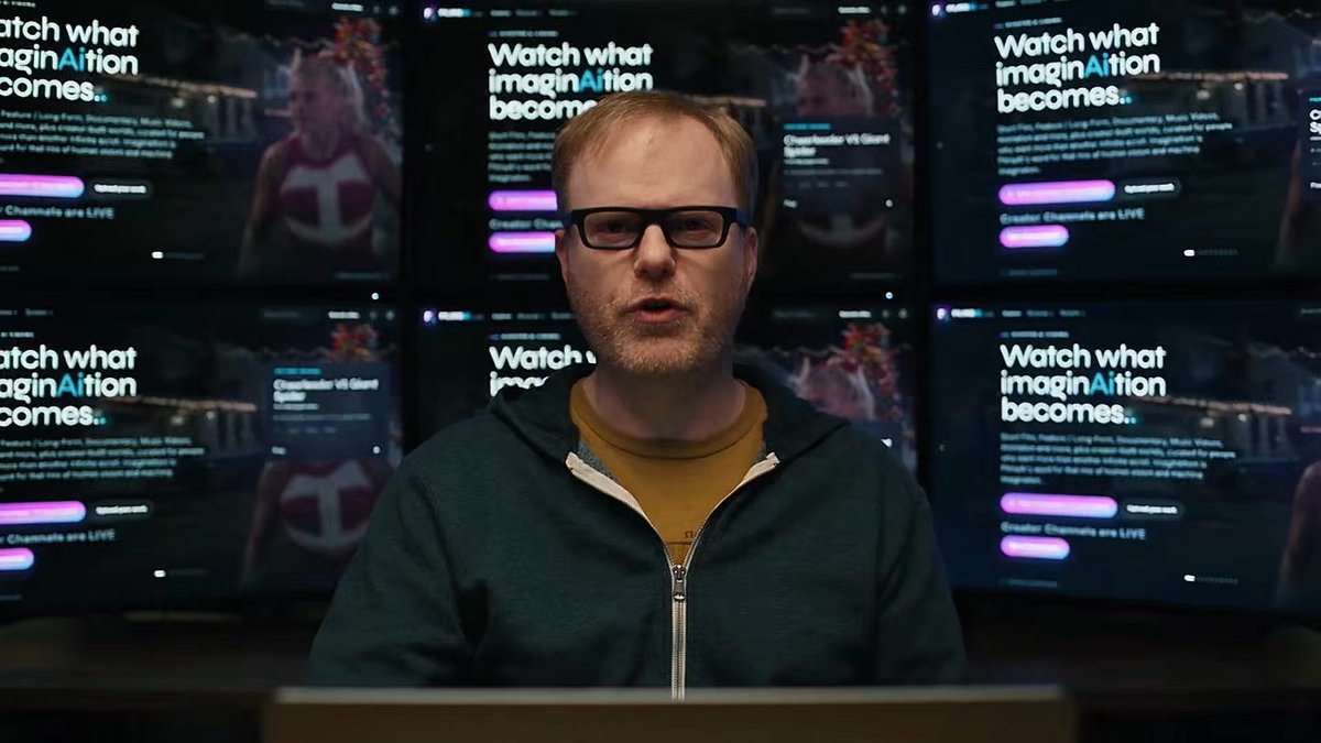 A man wearing glasses and a green hoodie stands in front of multiple monitors displaying the "Watch what imagination becomes.." web page with the text "Crosshair vs Giant Spider."