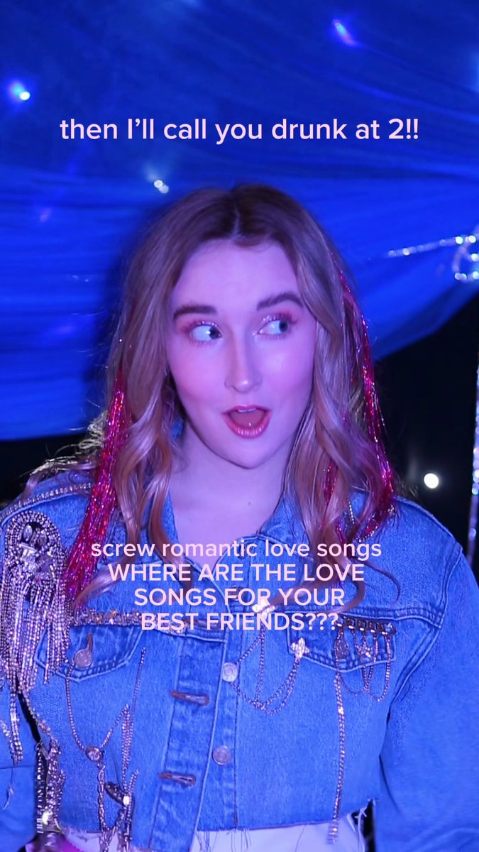 A woman with reddish hair and a denim jacket stands under blue stage lighting, with overlaid text reading 'then I'll call you drunk at 2!!' and 'screw romantic love songs WHERE ARE THE LOVE SONGS FOR YOUR BEST FRIENDS???'