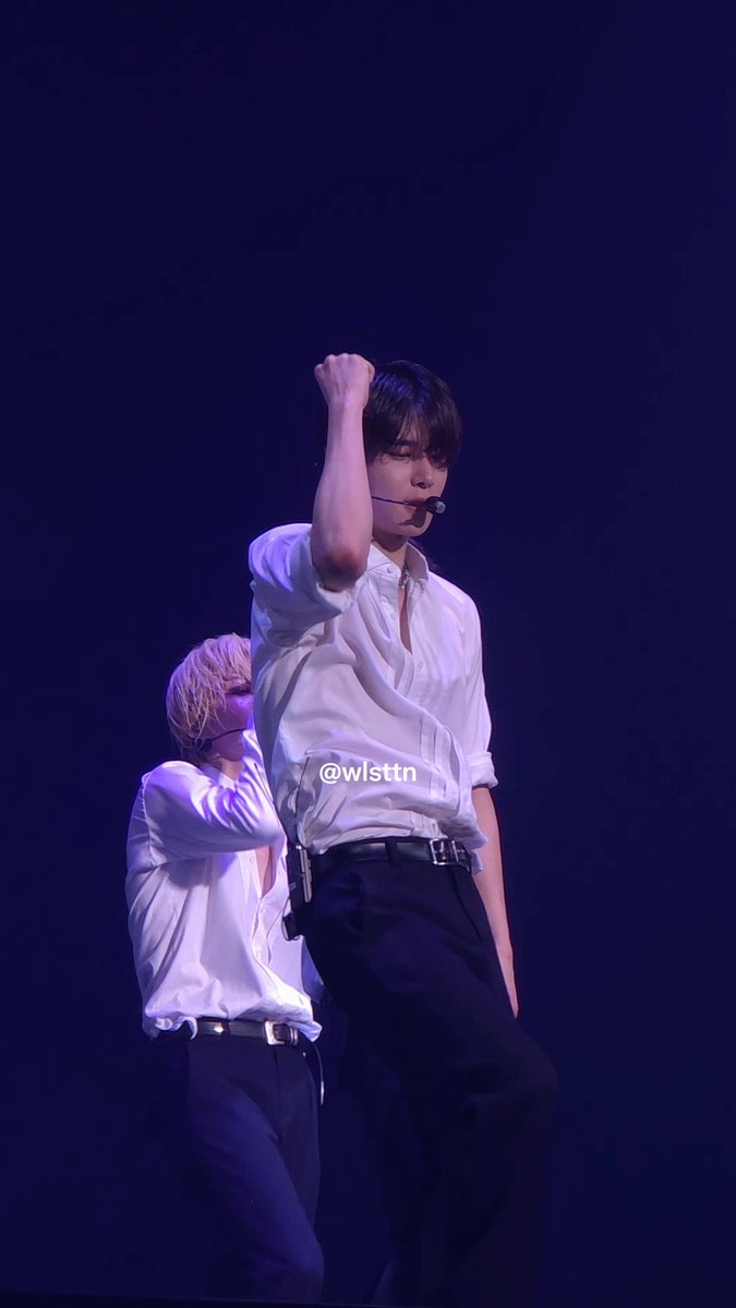 A concert performance photo with watermark @wlsttn showing a male performer in a white shirt raising his fist, with another performer in the background on a dark stage.