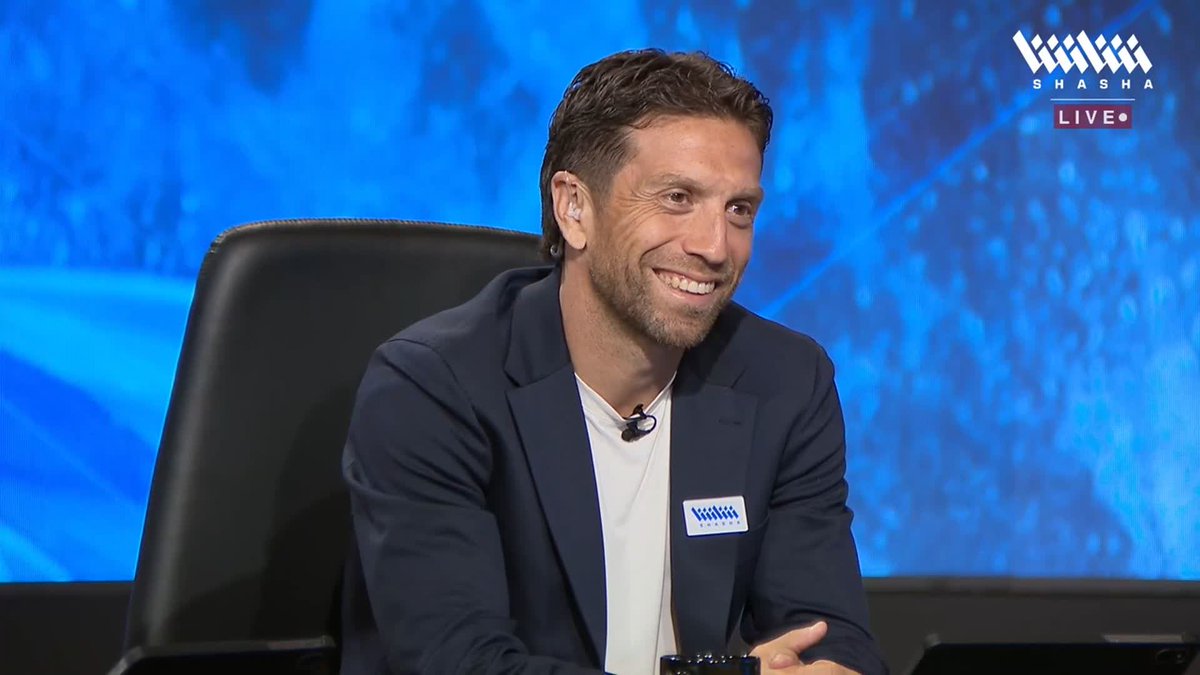 A smiling man in a navy blazer and white t-shirt sits in a studio chair against a blue backdrop, with a "Shasha Live" logo visible in the upper right corner and a "Shasha" name tag on his jacket, consistent with a sports analyst commenting on Messi's match against Benin on the Shasha app.