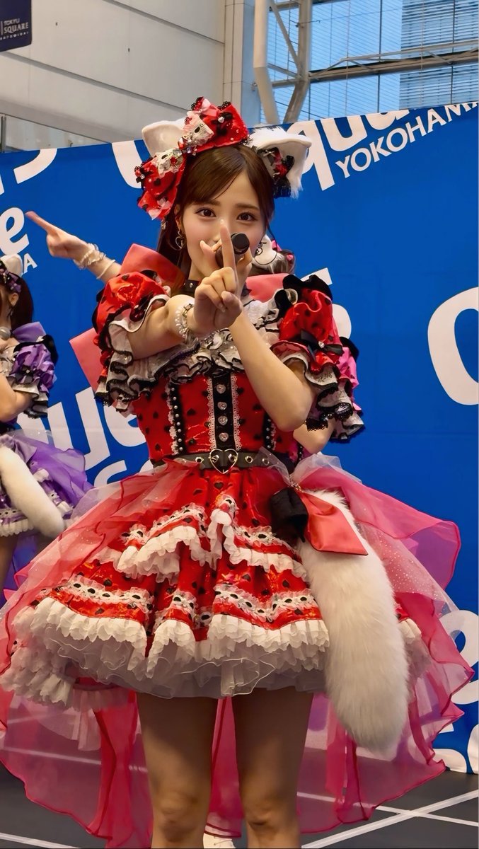A young woman in an elaborate red and black frilly idol costume with a large bow and headpiece, posing with peace signs in front of a blue 'YOKOHAMA' backdrop at what appears to be a cosplay or idol event.