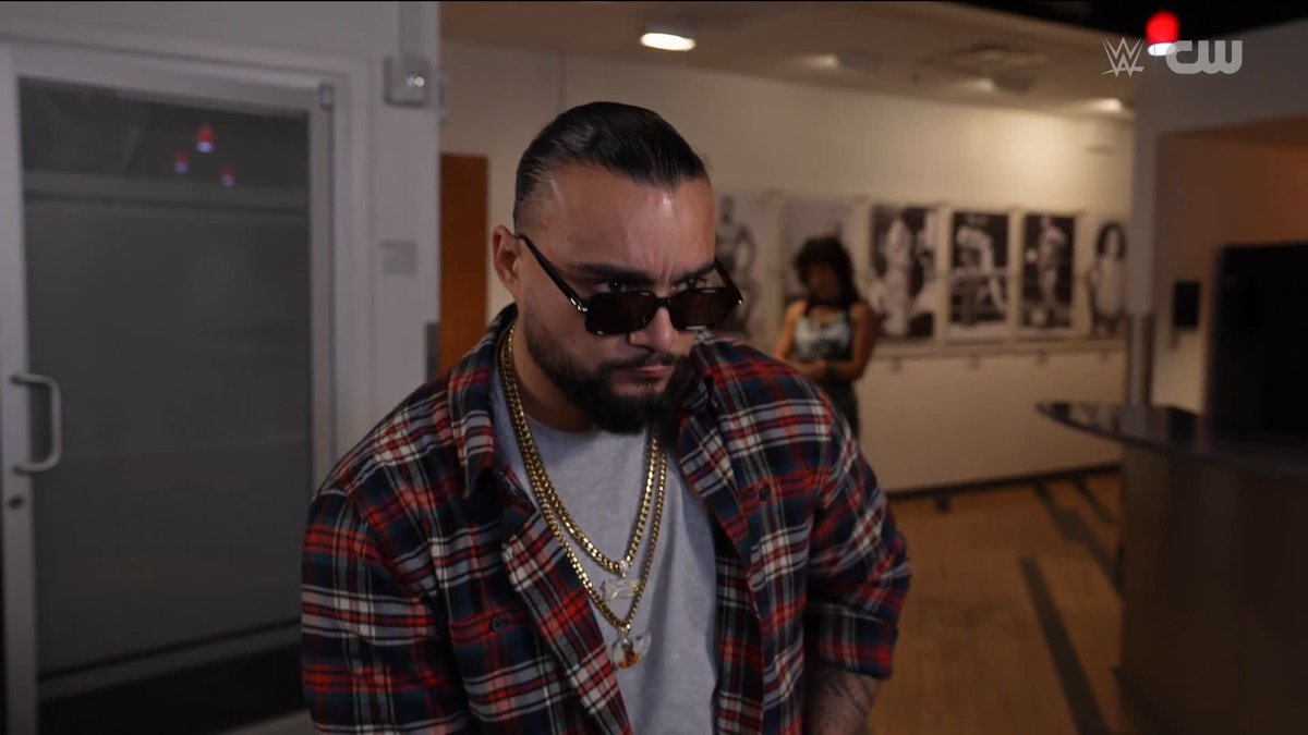 A bearded man wearing a plaid shirt, sunglasses, and gold chains stands in what appears to be a backstage area, with framed black-and-white photos visible on the wall behind him. The WWE and CW logos appear in the upper right corner, suggesting this is from a WWE-related production featuring Montana of NYPR.