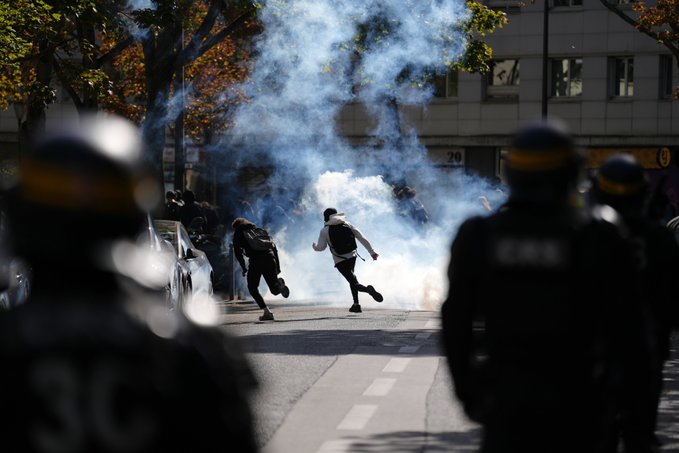 People run through clouds of tear gas on a street as riot police in helmets look on, with the word 'POLICE' visible on their uniforms.