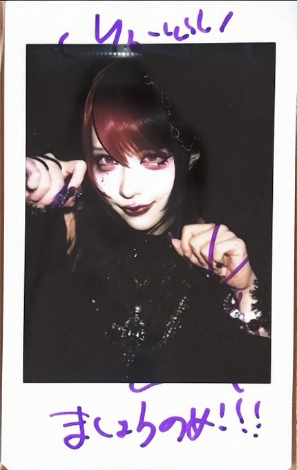 A polaroid photo of a person with red and black hair, pale makeup, and dark lipstick, dressed in gothic-style clothing with chains and spiked jewelry, featuring handwritten Japanese text and purple marker scribbles on the white border.