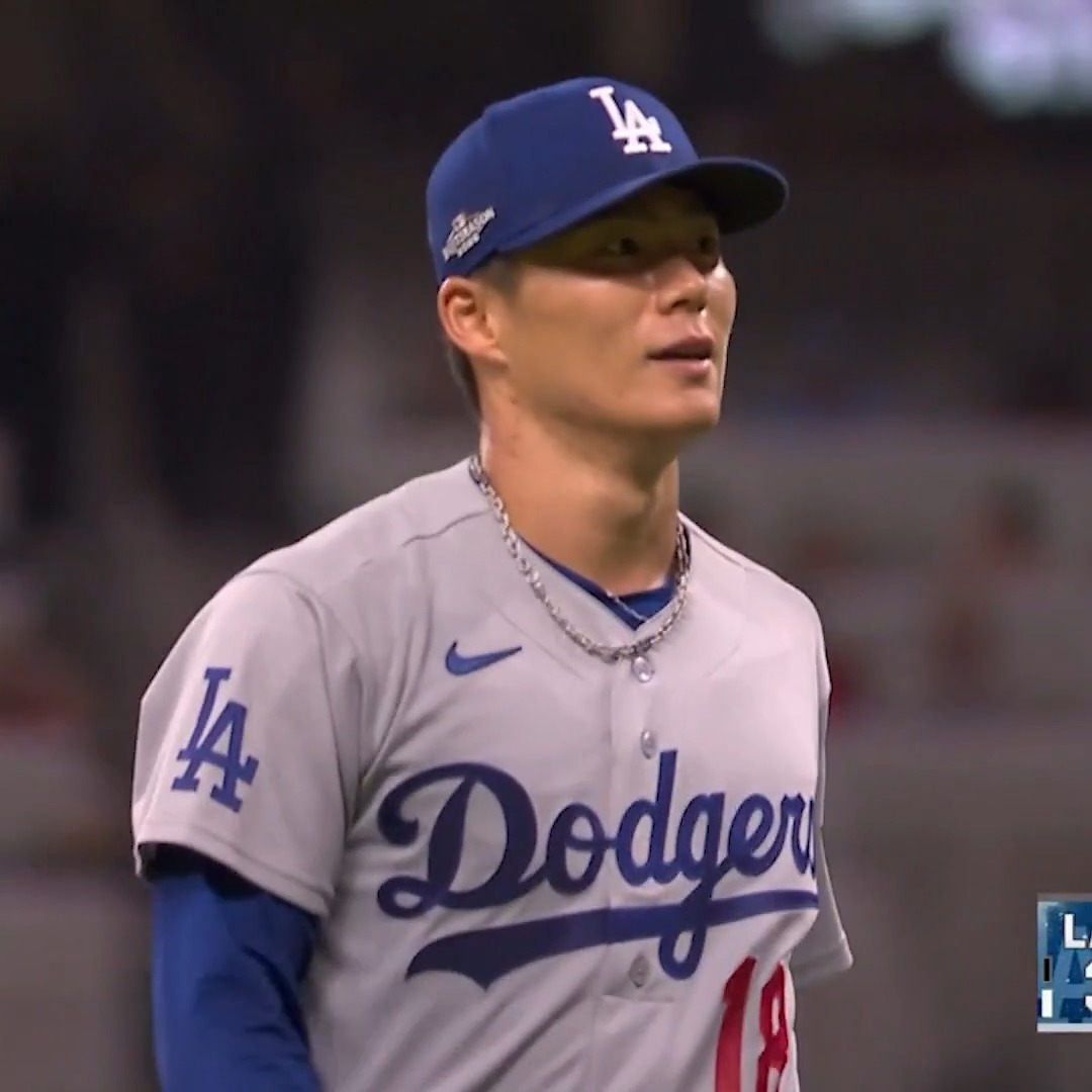 A baseball player wearing a Los Angeles Dodgers jersey and cap, with an 'LA' logo, standing in a stadium.