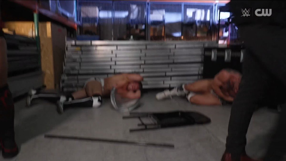 A wrestler from BirthRight lies sprawled on the mat after an attack, with an overturned folding chair nearby and another wrestler down on the right, while WWE and CW logos appear in the upper right corner. The image captures the aftermath of the surprise assault described in the caption, showing the chaotic scene in what appears to be a backstage area with metal barriers in the background.