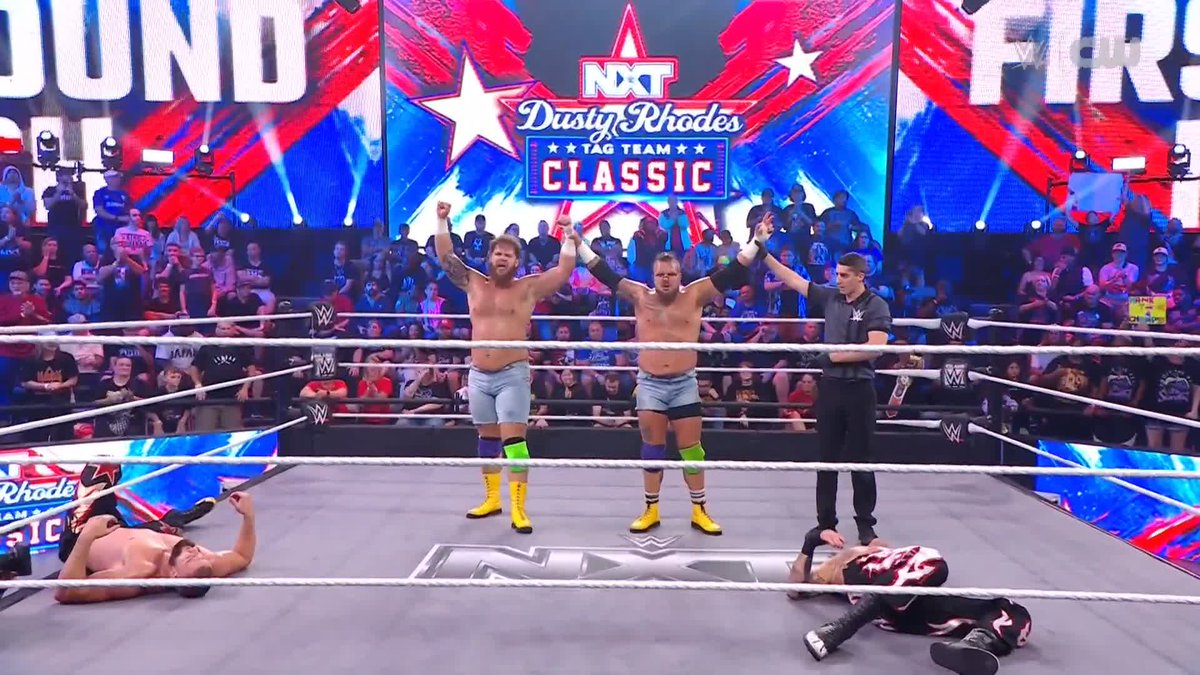 Two shirtless wrestlers in denim shorts and yellow boots stand victorious in the ring with their arms raised, while a referee raises one wrestler's hand amid two fallen opponents, under banners reading "NXT Dusty Rhodes Tag Team Classic" and "ROUND" — celebrating Hank and Tank's advancement.