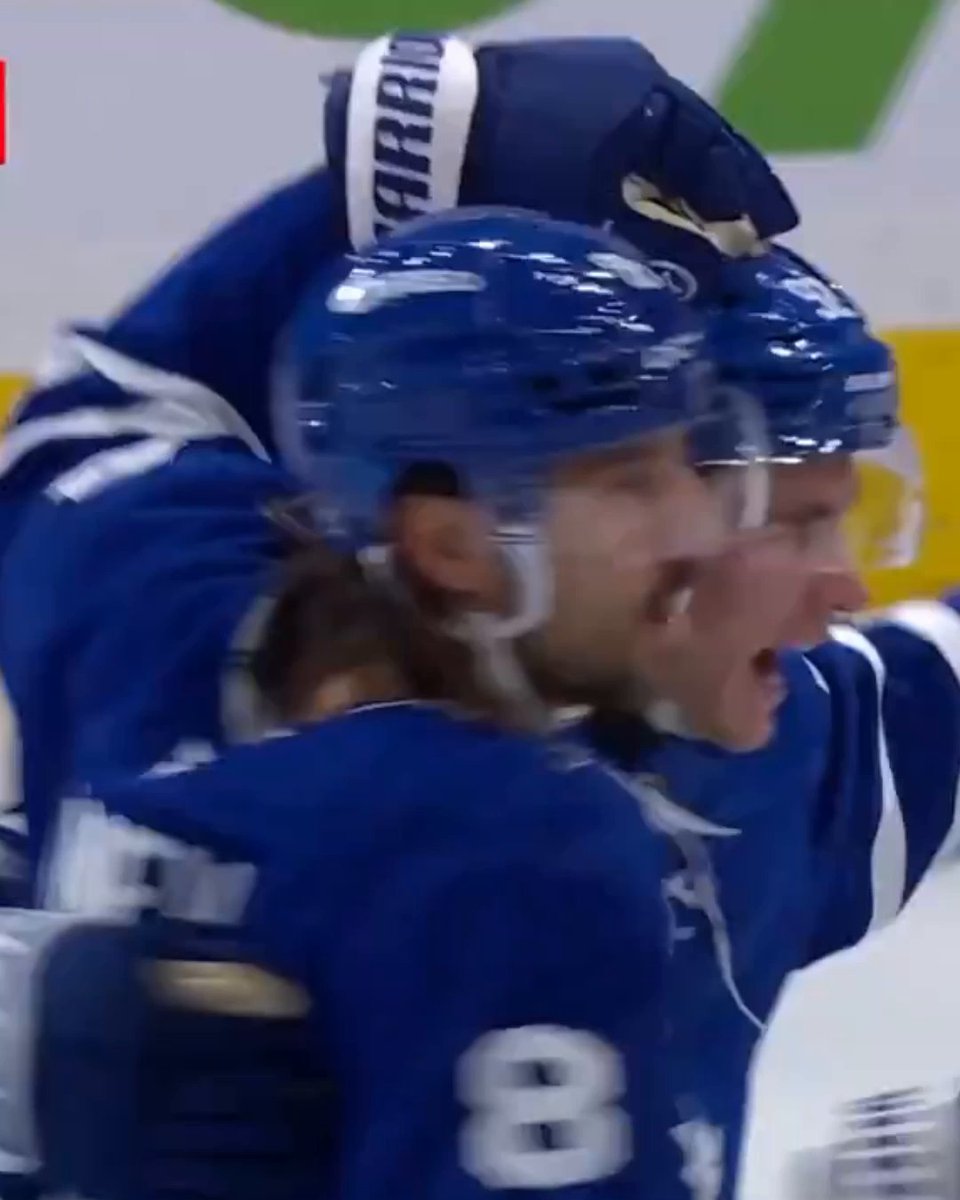 Tampa Bay Lightning players wearing blue jerseys and Warrior-branded helmets celebrate in an animated huddle, with the number 8 visible on one player's jersey, capturing the emotional aftermath of Gavin McKenna's first NHL goal assisted by Auston Matthews.