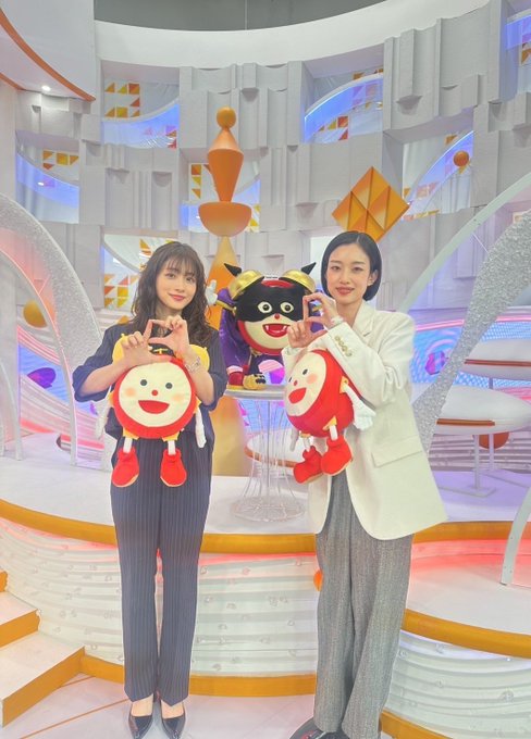 Two smiling women pose in a bright, modern game show set while holding plush toy mascots and forming heart shapes with their hands, with a costumed character visible behind them. The glossy, saturated colors match the comment's playful suggestion that the photo looks like it was taken straight from a steamer, giving it that over-processed, polished aesthetic typical of Japanese variety show graphics.
