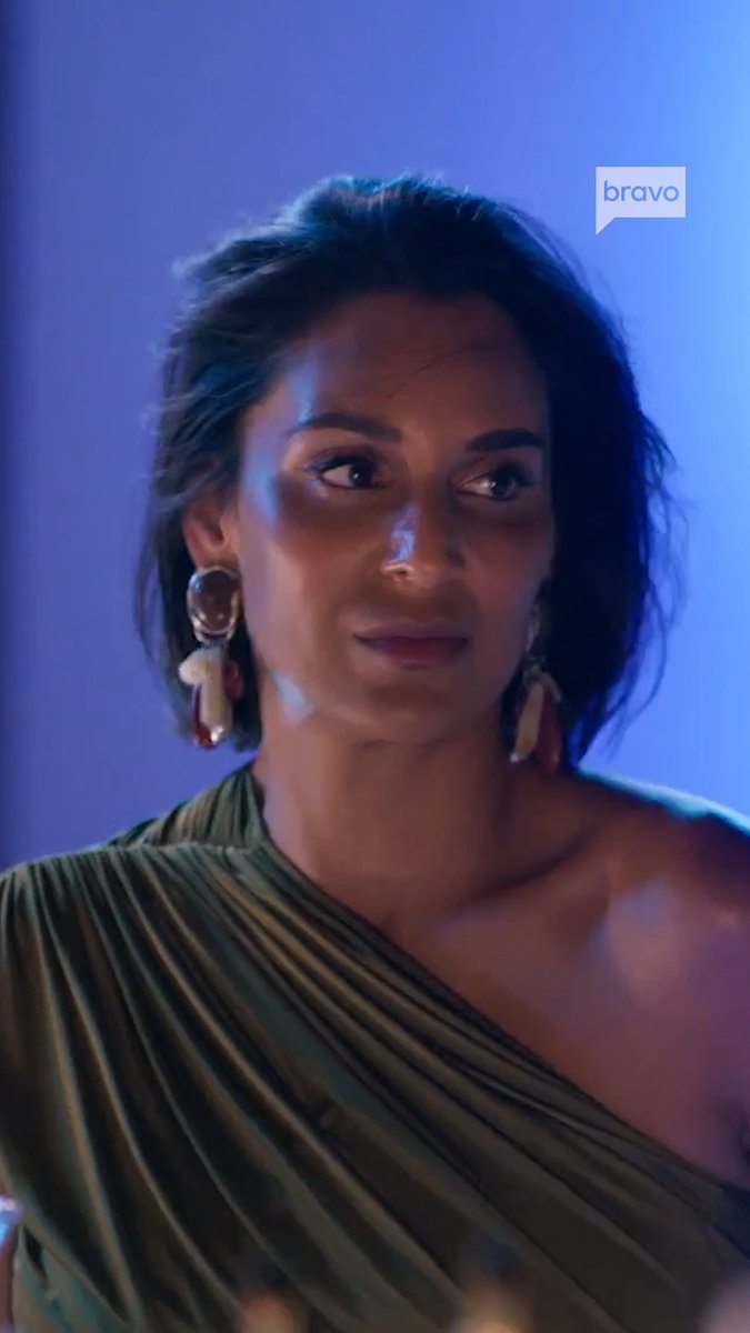 A woman in an olive green one-shoulder dress looks off to the side with a serious expression, while the "bravo" logo appears in the upper right corner. The dramatic blue lighting and her attentive gaze match the post's note that Daisy is asking questions about Moskito Island on #RHONY.