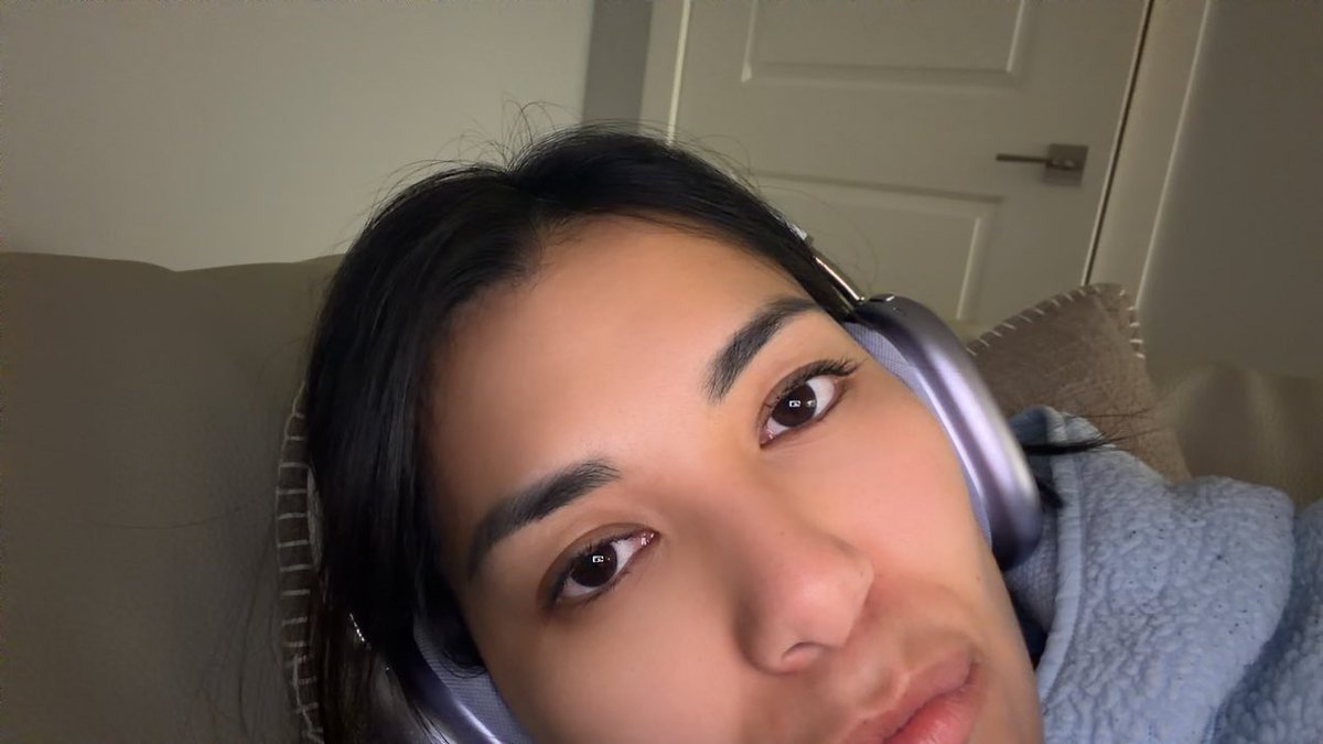 A close-up selfie shows a young woman with dark hair wearing rose-gold over-ear headphones and a light blue fuzzy sweater.