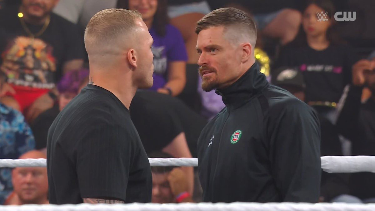 Two WWE wrestlers, one with short reddish-brown hair in a black t-shirt and another sporting a goatee and undercut in a black "UAT" jacket, stare each other down intensely inside the ring. The CW logo appears in the top right corner, suggesting a potential feud between the competitors.