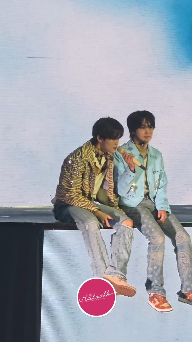 Two male performers sit on the edge of a stage; one wears a zebra-print jacket and ripped jeans, while the other wears a light blue jacket and holds a microphone. A watermark reads 'Haehyukku'.