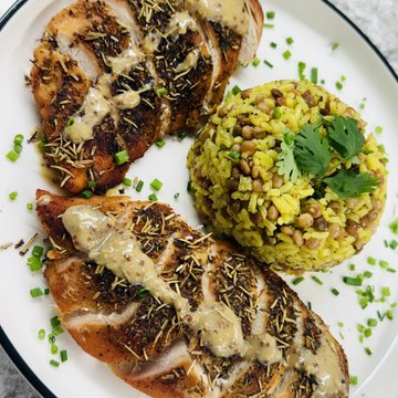 A plate of food featuring two seasoned grilled chicken breasts drizzled with sauce, served alongside a mound of yellow rice mixed with greens and garnished with cilantro.