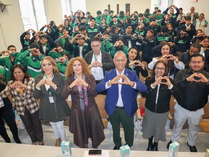 A large group of people in a tiered room forming heart shapes with their hands; adults in business attire stand in the front row, while students in green and navy tracksuits fill the rows behind them.